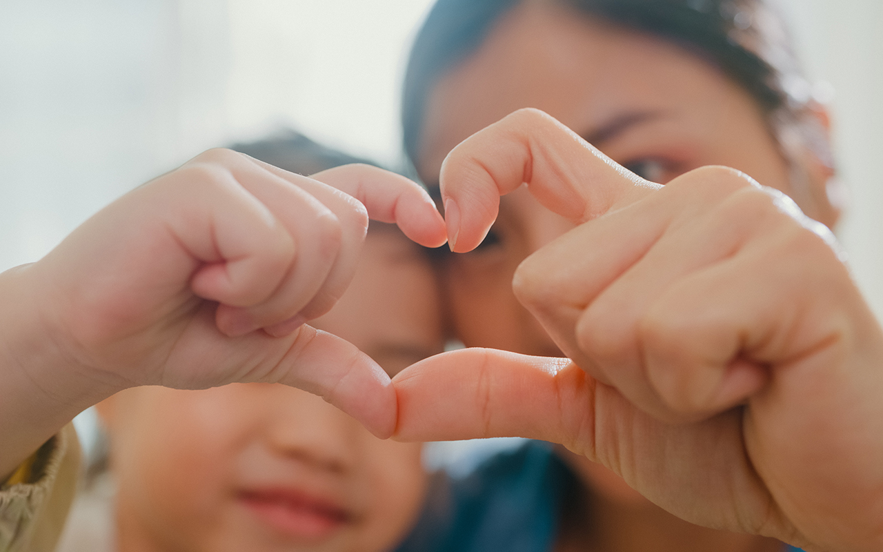 Closeup of young Asian family child making fingers heart shaped signs on sofa in living room at home. Love showing hand heart gesture, Family happy moment. income-tax-form-pen-finance-concept